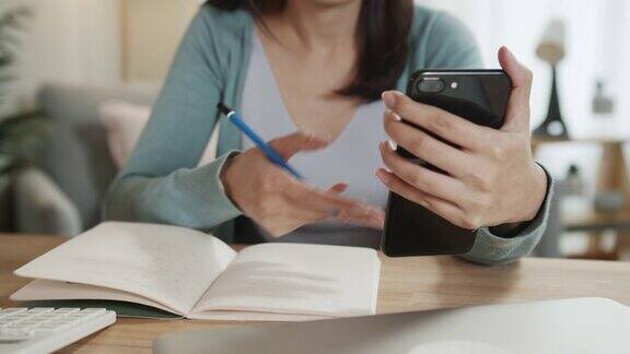 亚洲女性自由职业者手使用智能手机检查电子邮件阅读商业报告同时在笔记本上做笔记在家庭办公室工作