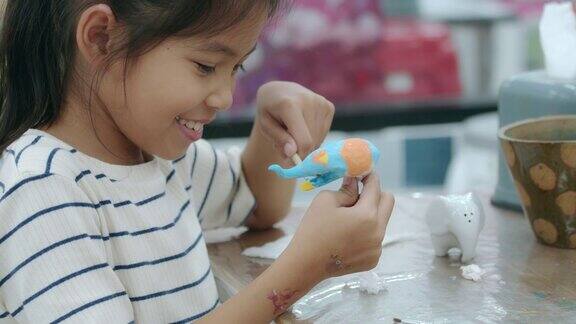 亚洲小女孩的慢镜头在小陶瓷象上兴致勃勃地画着油画孩子们在学校的艺术和手工艺创意活动班