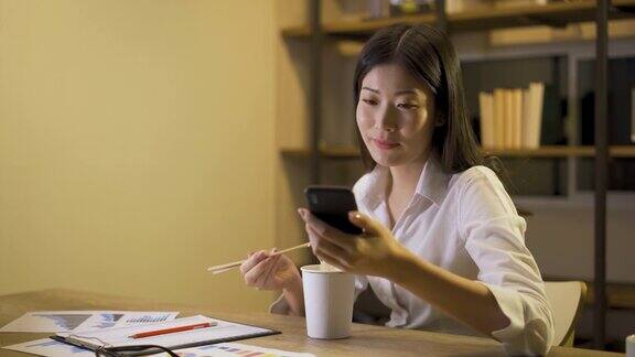 聪明的亚洲自由职业商业女性在家客厅用手机工作的同时吃着方便面