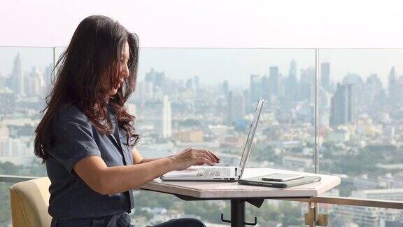 深思熟虑的亚洲妇女思考写文章的创意想法阅读电子邮件在办公室休息室的摩天大楼和模糊的背景城市在笔记本电脑努力工作与无线互联网