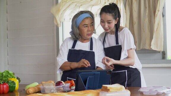 快乐的亚洲年轻的女儿和年长的母亲烹饪网上班在平板电脑上一起做新鲜的蔬菜食物在厨房在家里