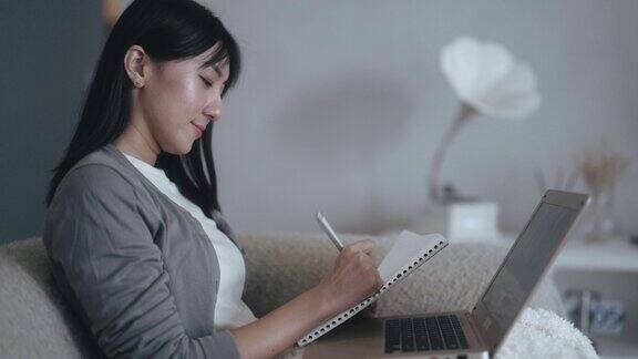 亚洲华人妇女在家工作坐在沙发上用笔记本电脑写笔记