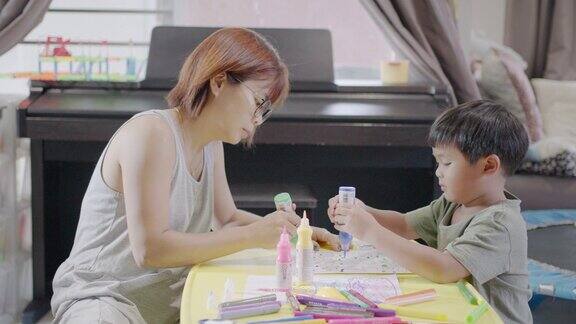 快乐的亚洲男孩与他的母亲在家里的客厅画水彩画