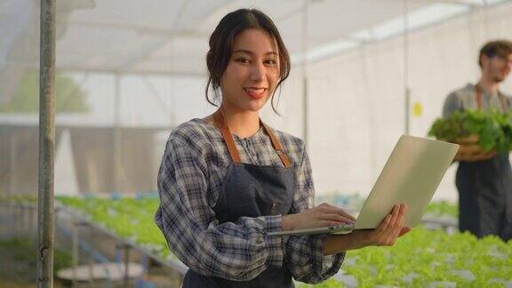 微笑的亚洲女性年轻的农民走到检查沙拉蔬菜在一个水培农场种植在温室用她的手和爱做在笔记本电脑上为质量控制在我们的小型农业农场在日落下