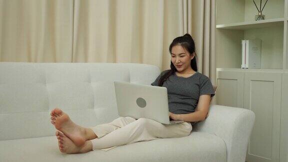 亚洲妇女坐在沙发上使用笔记本电脑在家里工作