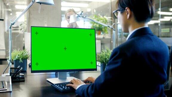 在办公室里商人在他的办公桌上用一台模拟的绿色屏幕的个人电脑工作同事走进办公室并在她的办公桌前发生