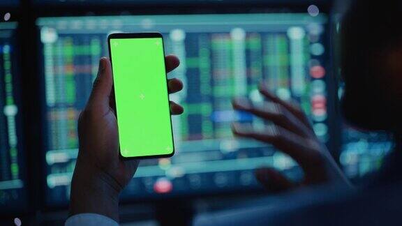 金融分析师使用智能手机与绿色屏幕色度键模拟模板和工作在多显示器工作站与股票图表商人晚上在投资银行工作