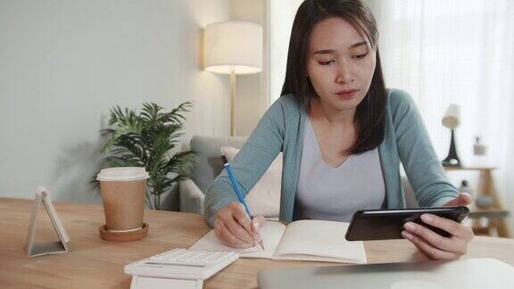 亚洲女性自由职业者使用智能手机检查电子邮件阅读商业报告同时在笔记本上做笔记在家办公