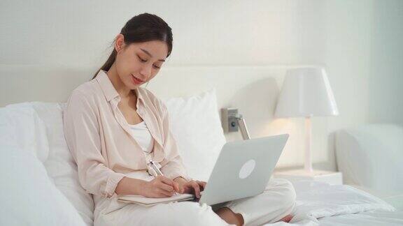 亚洲女商人写笔记本和使用技术笔记本工作在线学习在床上