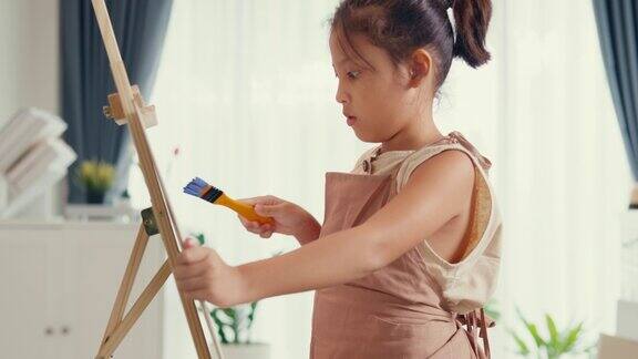 特写亚洲蹒跚学步的女孩与围裙坐在沙发椅前的油画画布与调色板使用油漆刷专注于绘画抽象周末在家