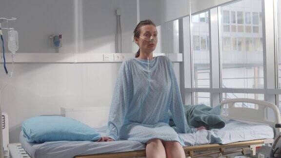 年轻的女病人带着鼻插管坐在病床上在病房的窗帘后面呼吸