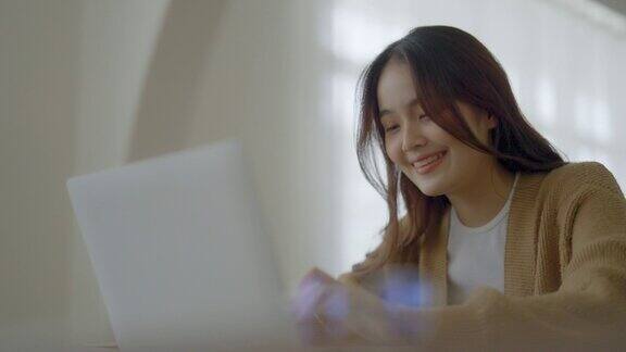 微笑的亚洲年轻女性在家庭办公室的笔记本电脑上工作年轻的亚洲学生使用计算机远程学习虚拟培训电子学习观看在线教育在家里的网络研讨会