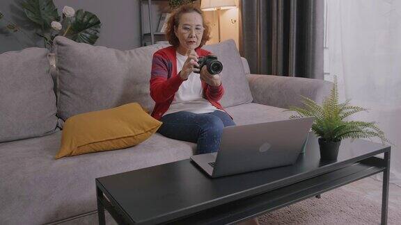 广角拍摄和手持:迷人的亚洲高级女性通过笔记本电脑学习在线摄影课坐在家里客厅的沙发上和她的老师学习如何用视频通话技术使用数码相机