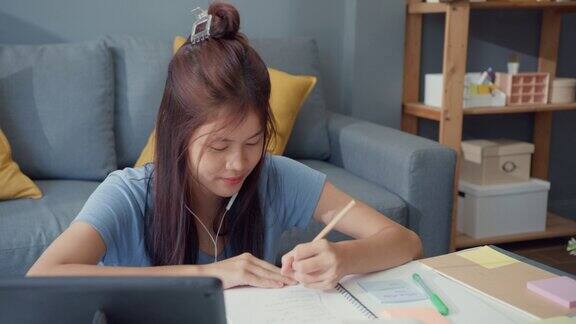 年轻的亚洲女孩青少年戴着休闲耳机使用数码平板电脑在线学习写演讲笔记本在客厅在家里隔离教育在线电子学习冠状病毒大流行概念