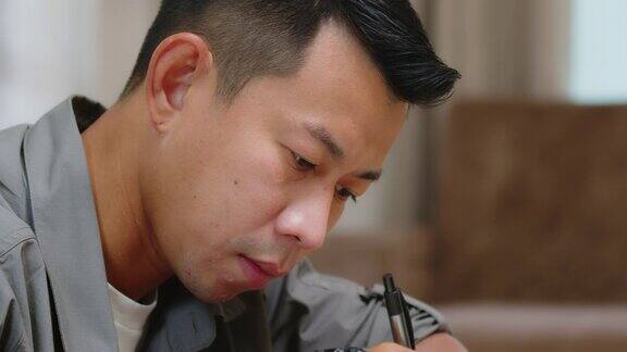 近距离的亚洲学生男子的脸握笔和写在笔记本上而学习在线在客厅在家里