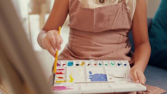 特写亚洲蹒跚学步的女孩与围裙坐在沙发椅前的油画画布与调色板使用油漆刷专注于绘画抽象周末在家