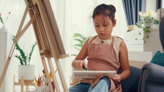 亚洲学步女孩用围裙坐沙发椅子前面的油画画布把丙烯酸色在调色板上使用调色板刀混合颜色和做抽象艺术在客厅家里