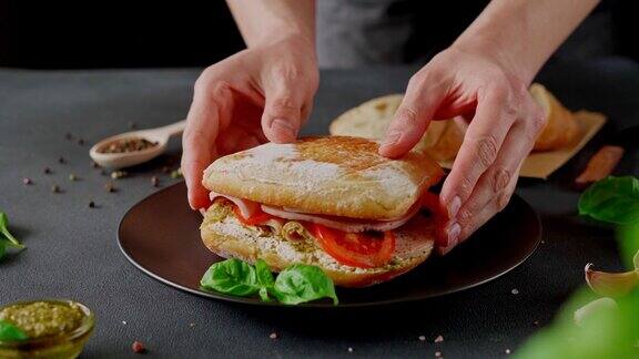 男人的手准备恰巴塔三明治与火腿罗勒香蒜沙司西红柿在黑色背景食品的背景