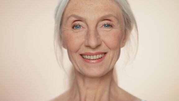 老年妇女的肖像看着相机微笑自然灰色头发蓝眼睛的老妇人优雅的老年概念的护肤品美容化妆品产品与孤立的米色背景