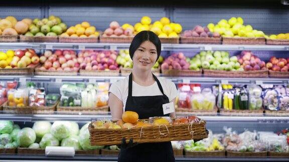 迷人的年轻亚洲女工人肖像站在超市货架水果的背景看着相机和微笑贸易、商业和人的观念