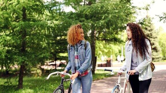 快乐的游客美丽的女人在美丽的绿色公园里骑着自行车和背包聊天自行车旅游千禧一代和人们