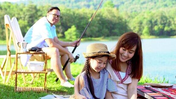 4K快乐的年轻亚洲家庭享受露营在湖两位父母和可爱的小女孩一家人坐在帐篷前的露营椅上钓鱼和玩得一起在假期