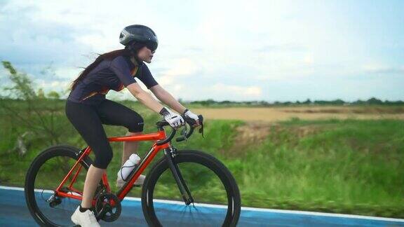 侧面跟踪亚洲女子橙色自行车戴着防护头盔锻炼训练快速骑在城市外的道路上