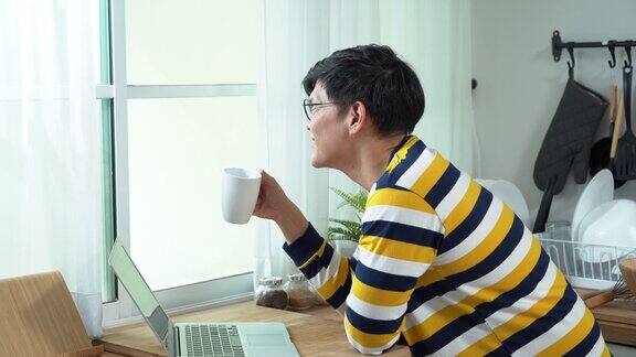 亚洲商人用笔记本电脑喝着咖啡在厨房里看着窗外在早晨的阳光下在家工作