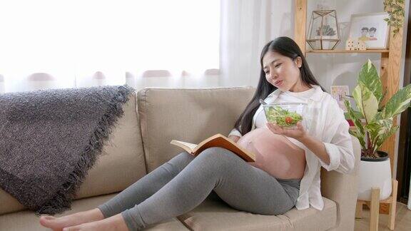 快乐的亚洲孕妇在家里的沙发上吃沙拉和看书怀孕期间多吃不同的蔬菜和放松健康的怀孕理念