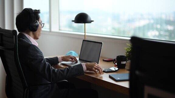 一位亚洲华人中年商人在办公室用耳机和笔记本电脑与同事和商业伙伴进行视频会议