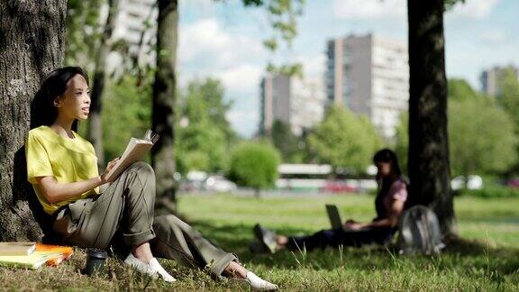 亚洲女学生坐在树下倚着树干在公园看书又一个大学女生在后台用笔记本打字