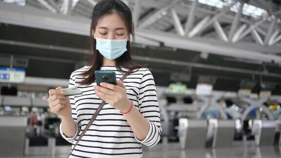 亚洲女性戴着口罩在机场使用移动支付预订航班因为Covid-19大流行她用智能手机预订和预订航班