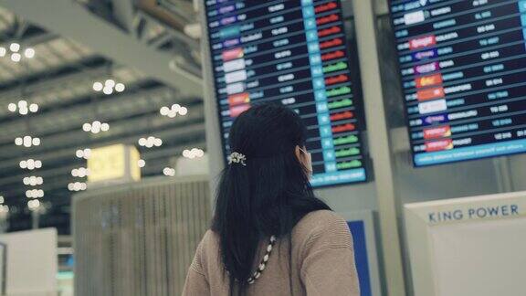 一名亚洲妇女戴着外科口罩在机场候机楼检查航班时刻表为了她的出差