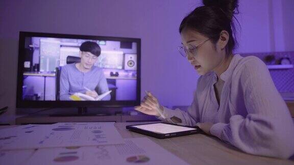 年轻的亚洲女性在办公室工作到深夜时用笔记本电脑进行视频通话与男性财务顾问学习