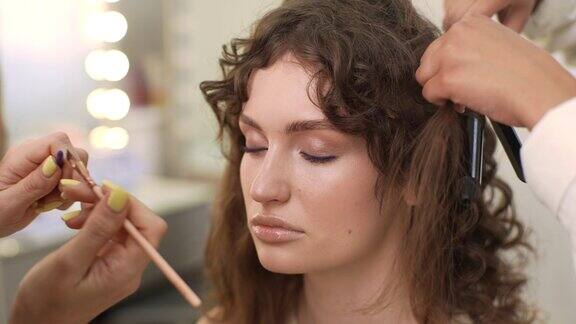 不知名的化妆师在年轻女子脸上画眼线的特写美发师用卷发钳为长发女士做发型