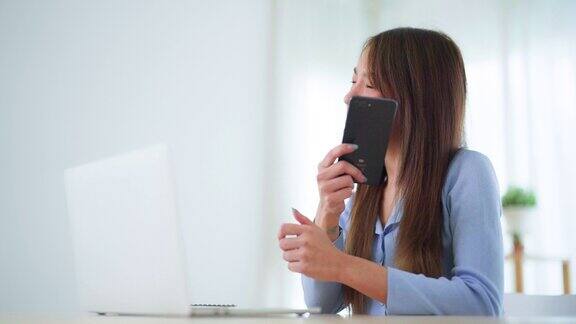 年轻的亚洲女子在家里客厅的桌子上用笔记本电脑工作时一边说着手机