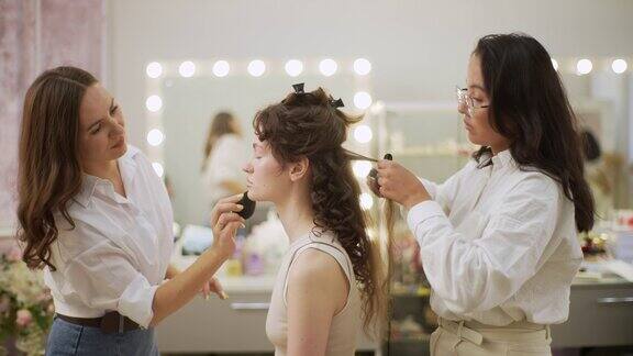 在美容院迷人的年轻女子正在用4只手化妆和做发型化妆师用海绵涂抹面部粉底