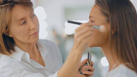 专业化妆师用刷子刷粉、化妆一个漂亮的女孩坐在美容院的一面大镜子前肖像的观点