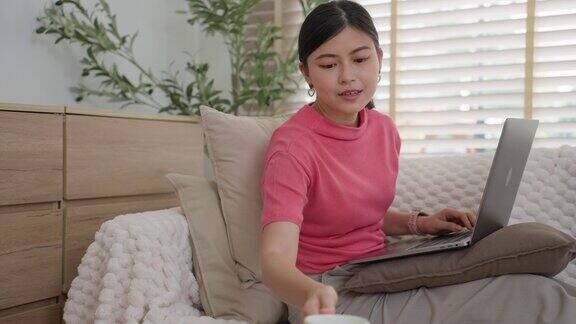 一个年轻的亚洲女人穿着休闲服坐在沙发上在房子里有百叶窗的窗户旁工作