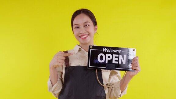 年轻的亚洲咖啡师女士穿着围裙举着黄色背景的开业招牌服务员或企业家兴高采烈地展示着招牌小企业或初创企业服务员或员工