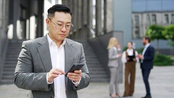 英俊的亚洲商人站在现代办公大楼附近使用手机户外