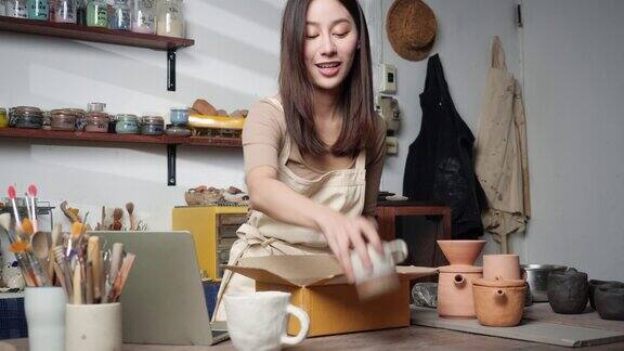 年轻的女士在把产品送到顾客面前检查它是否由粘土制成陶瓷美丽的女人包装陶瓷马克杯在盒子里女人使用笔记本电脑商业、技术和物流
