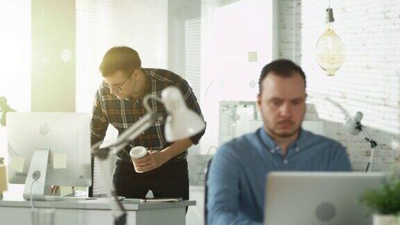 一名男子走进办公室坐在工作场所喝咖啡开始在个人电脑上工作在前景上一个人坐在办公桌前用笔记本电脑工作