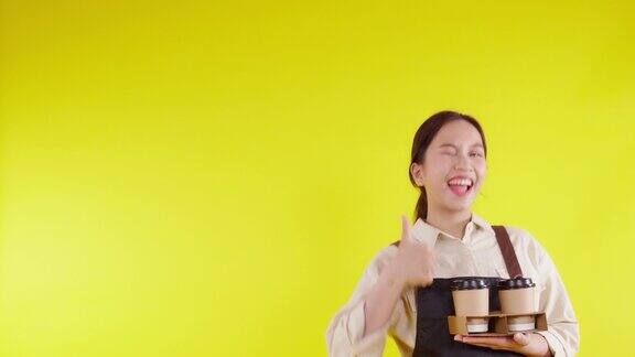 肖像年轻的亚洲咖啡师女士穿着围裙拿着咖啡杯呈现在黄色背景上服务员或企业家愉快地展示并竖起大拇指小企业或初创企业
