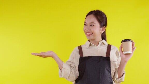 肖像年轻的亚洲咖啡师女士穿着围裙拿着咖啡杯呈现在黄色背景上服务员愉快地展示并竖起大拇指小生意或创业咖啡馆服务员