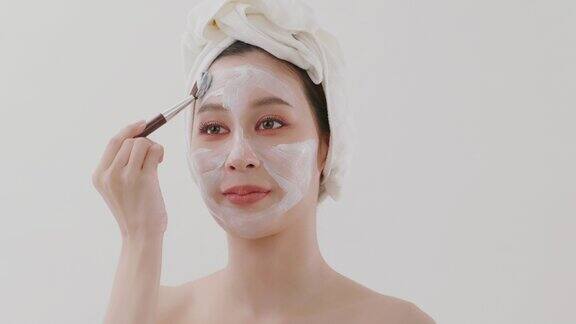 慢动作美丽的亚洲年轻女子与毛巾在她的头上应用粘土面膜的一半她的脸使用化妆刷和镜子