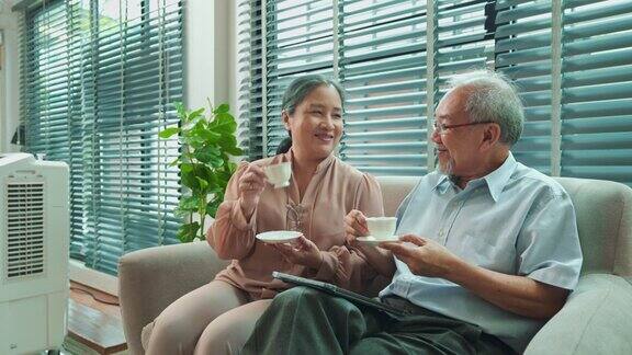 退休的亚洲老人坐在沙发上用平板电脑老妇人给他端来一杯茶