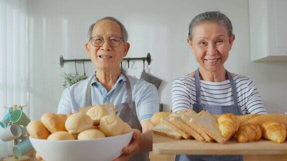 亚洲老年夫妇拿着一碗面包看着镜头的肖像上了年纪的成熟男女系上围裙在家里花时间做饭做早餐保健饮食与健康食品理念
