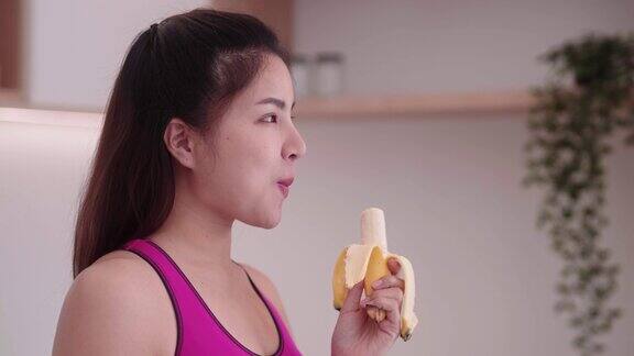 运动的年轻亚洲女性穿着运动服在厨房内吃香蕉享受吃水果的饮食和健康的运动减肥概念