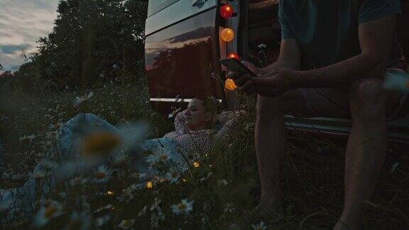 黄昏时分一名男子在草地上玩智能手机而一名女子躺在一辆露营车旁